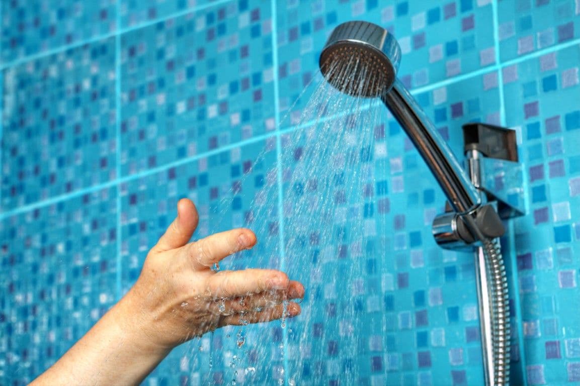 An outstretched hand underneath a running shower head in a blue tile shower stall.